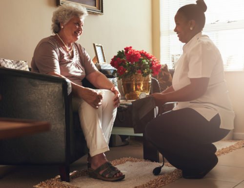 Senior woman sitting on a chair at home with female caregiver holding blood pressure gauge.  Female nurse visiting senior patient for checking blood pressure.